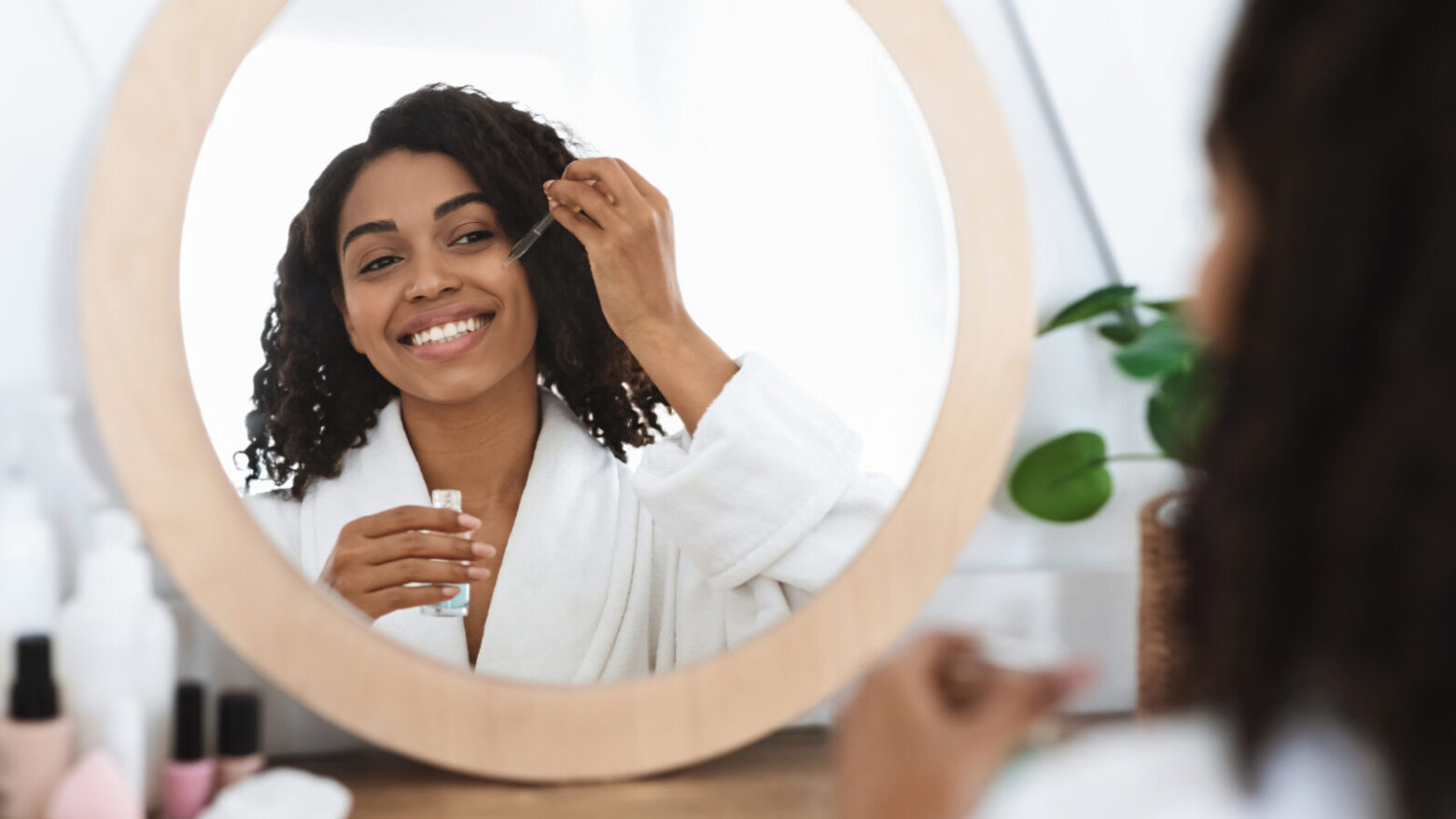 A woman sitting in front of a vanity mirror, applying a dropper of oil to her face.