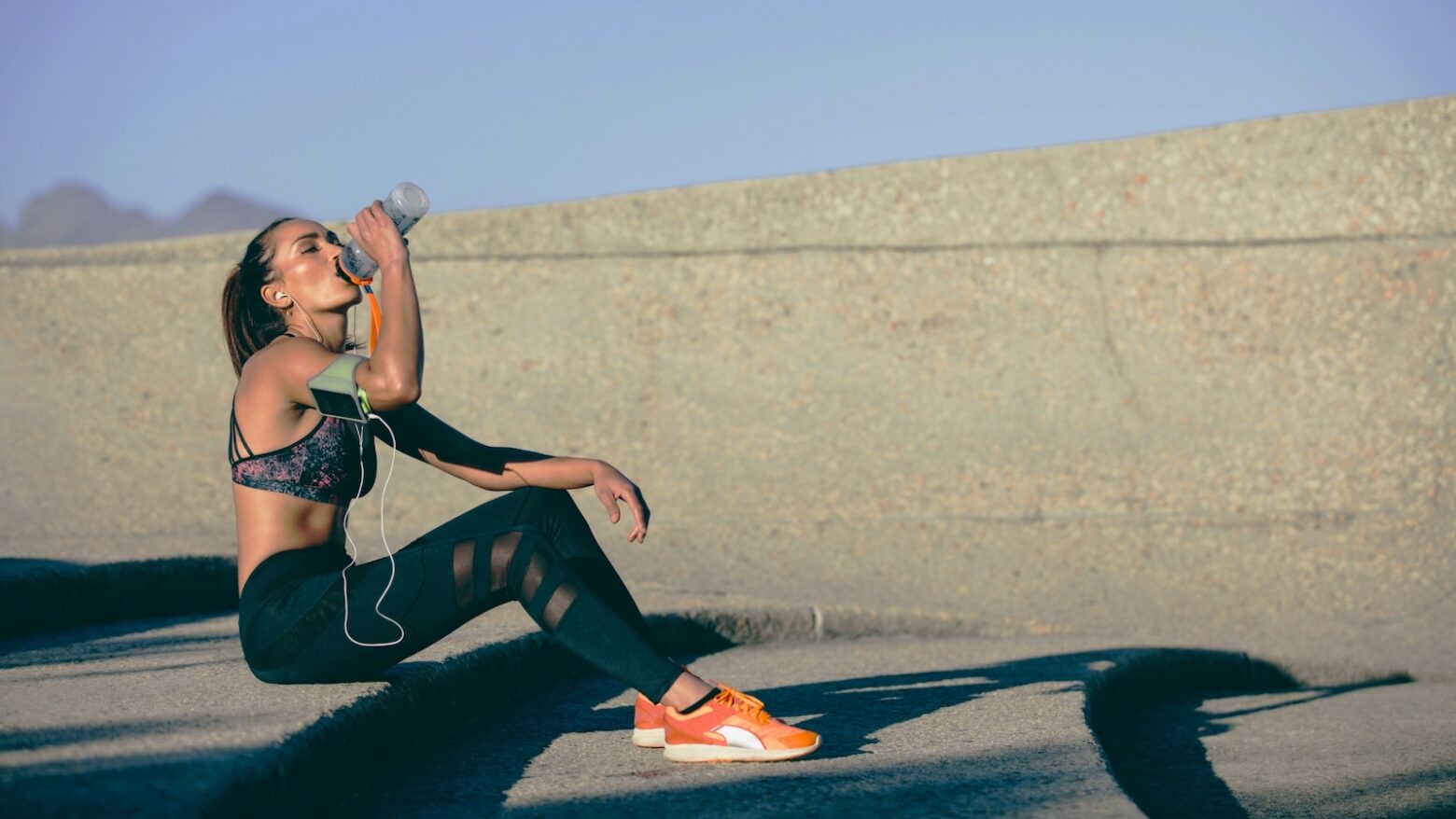 A woman in running shoes and related running gear drinks water on a step.