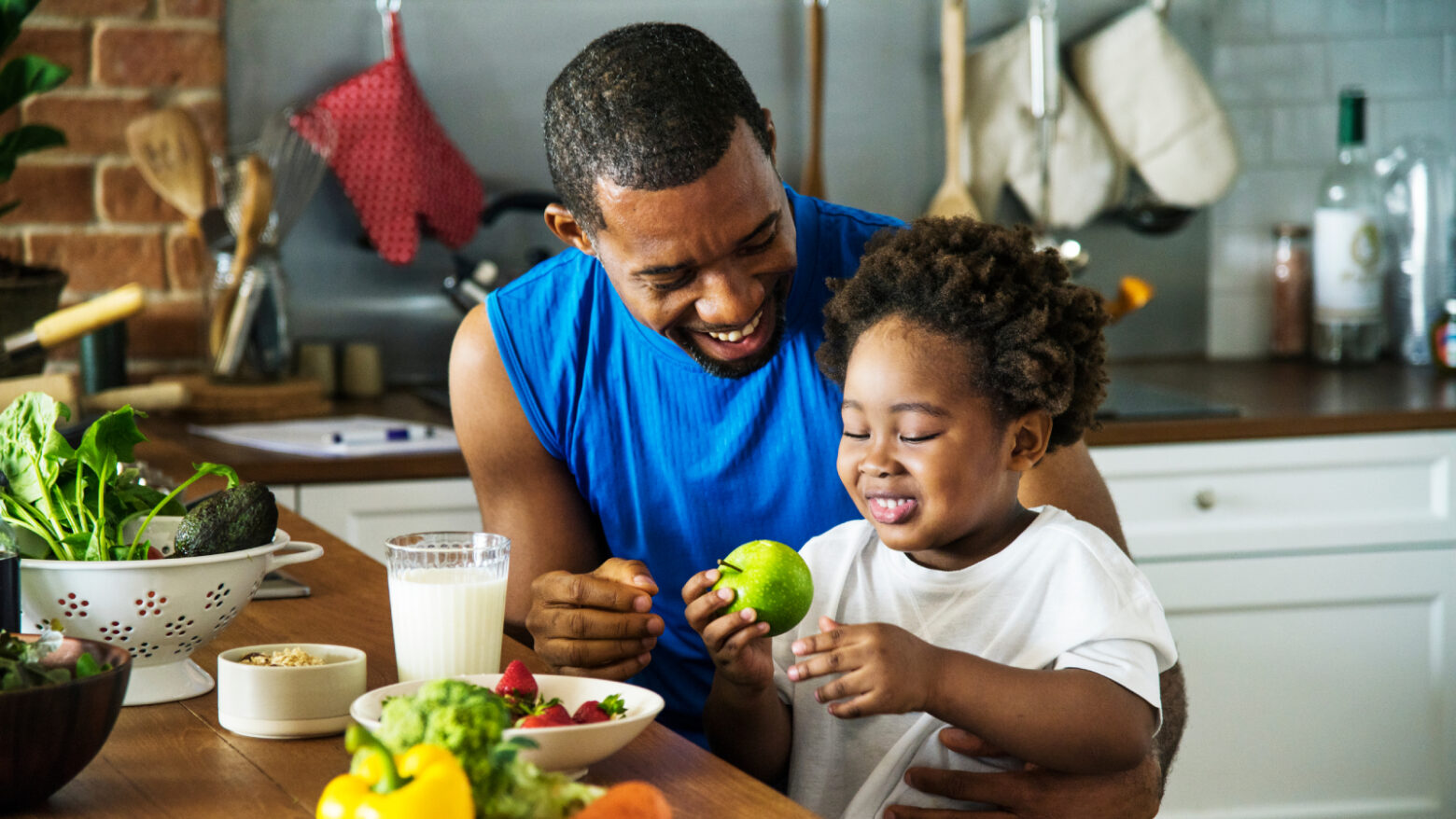 A father offers his child an apple while both are having lunch in the kitchen