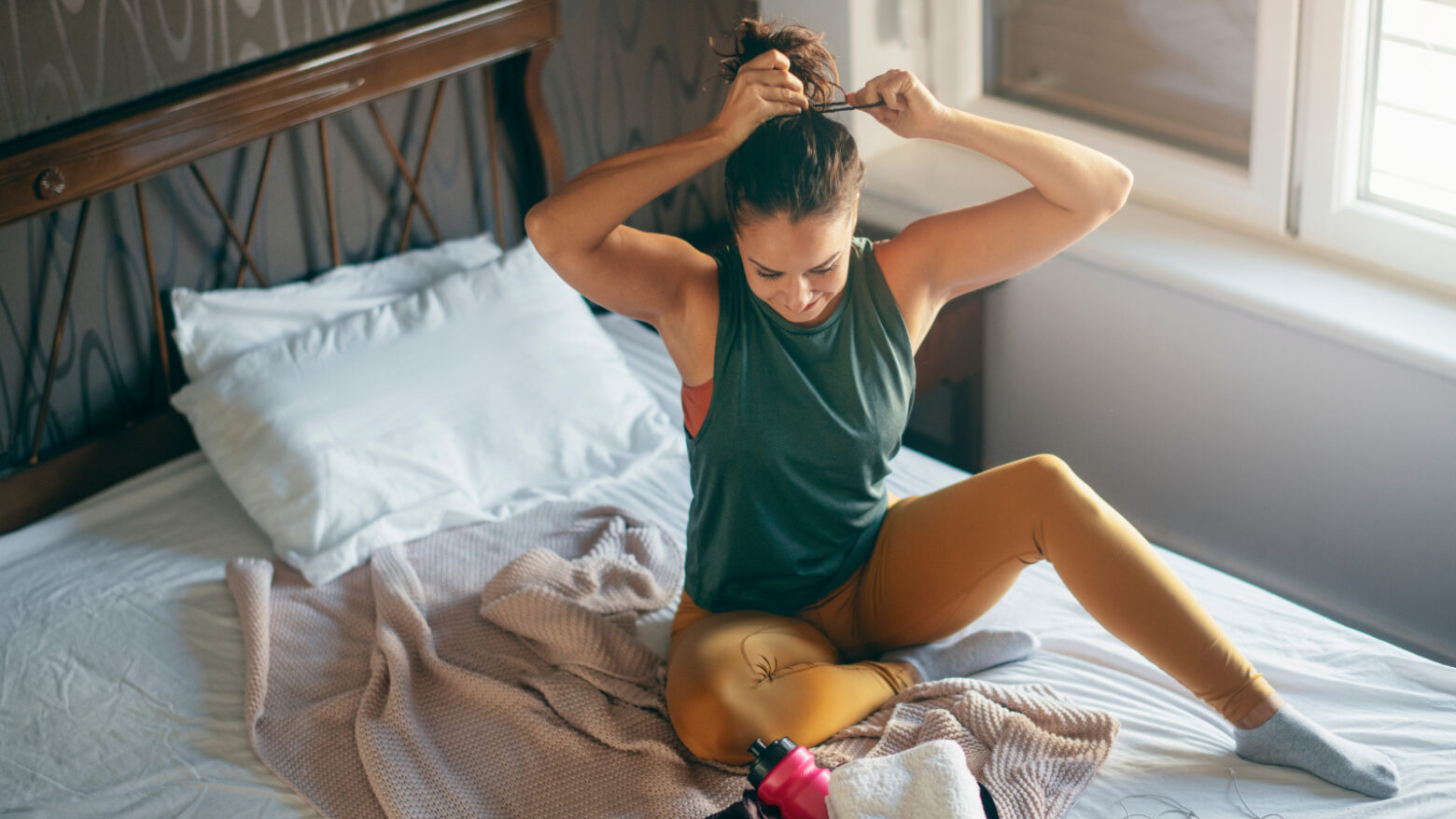 A woman ties up her hair while sitting on her bed in workout clothes