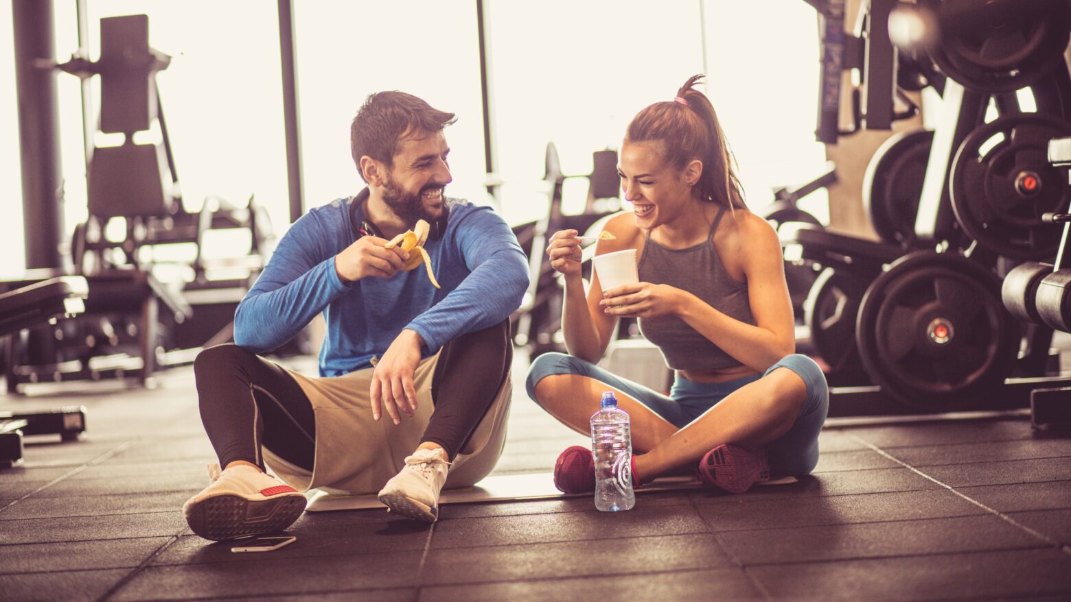 A happy couple sits on the floor of a gym while snacking on food