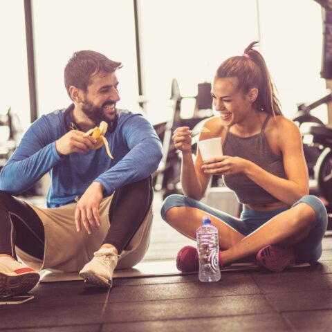 A happy couple sits on the floor of a gym while snacking on food