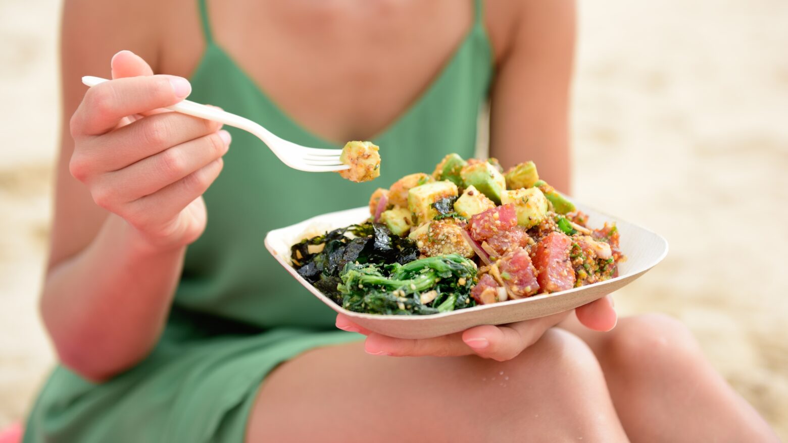 Close up of a woman eating a poke bowl at the beach.