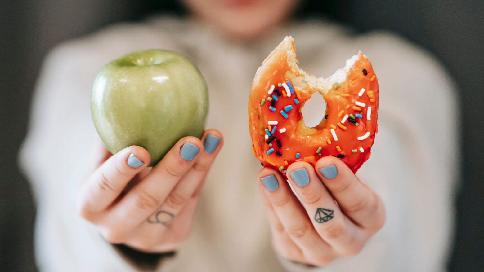 Close-up of a woman holding up a green apple and an orange-frosted donut with a bite taken out of it