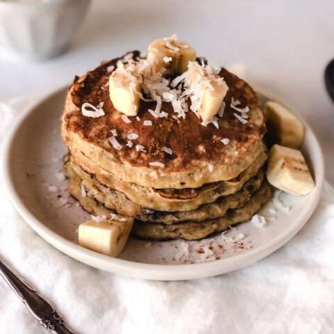 From above, four banana pancakes on a white plate garnished with shredded coconut and slices of bananas, next to a sliver fork.