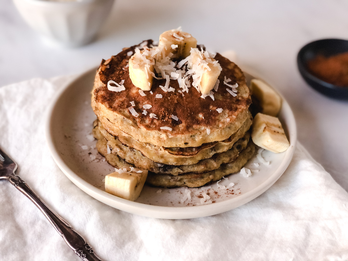 From above, four banana pancakes on a white plate garnished with shredded coconut and slices of bananas, next to a sliver fork.