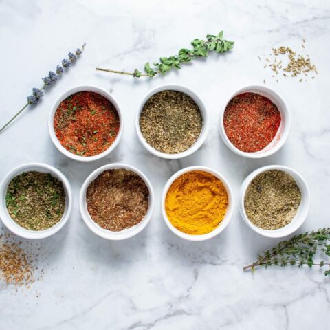 From above, seven small white bowls containing different spice blends with herbs and seeds surrounding them.