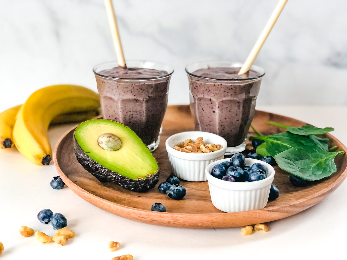 Two glasses of blueberry smoothie on a wooden platter with bananas, spinach leaves, half an avocado, and two ramekins of blueberries and walnuts