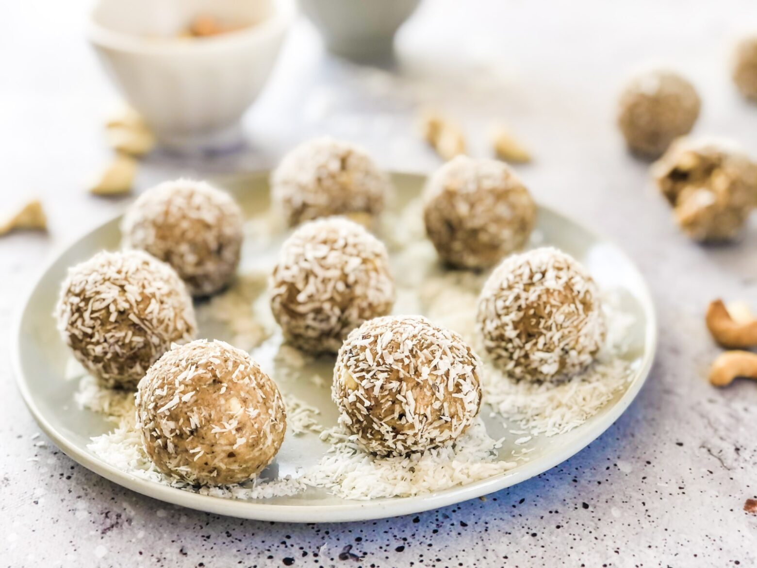 A white plate of coconut and cashew butter energy balls, garnished with shredded coconut surrounded by out-of-focus small bowls and ingredients.