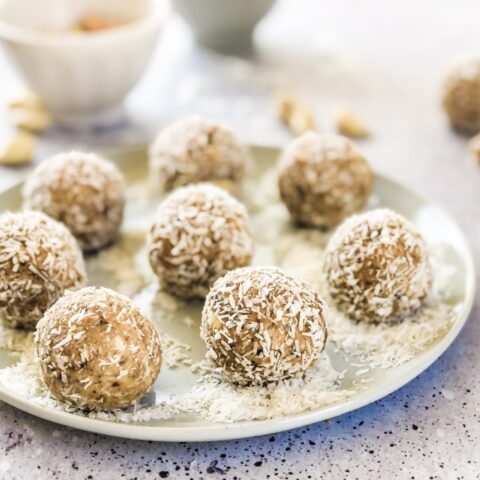 A white plate of coconut and cashew butter energy balls, garnished with shredded coconut surrounded by out-of-focus small bowls and ingredients.