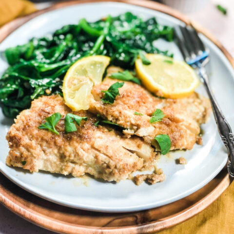 A plate of baked cod garnished with chopped fresh parsley and two lemon slices, with sautéed spinach and a silver fork, placed on a wooden charger plate.