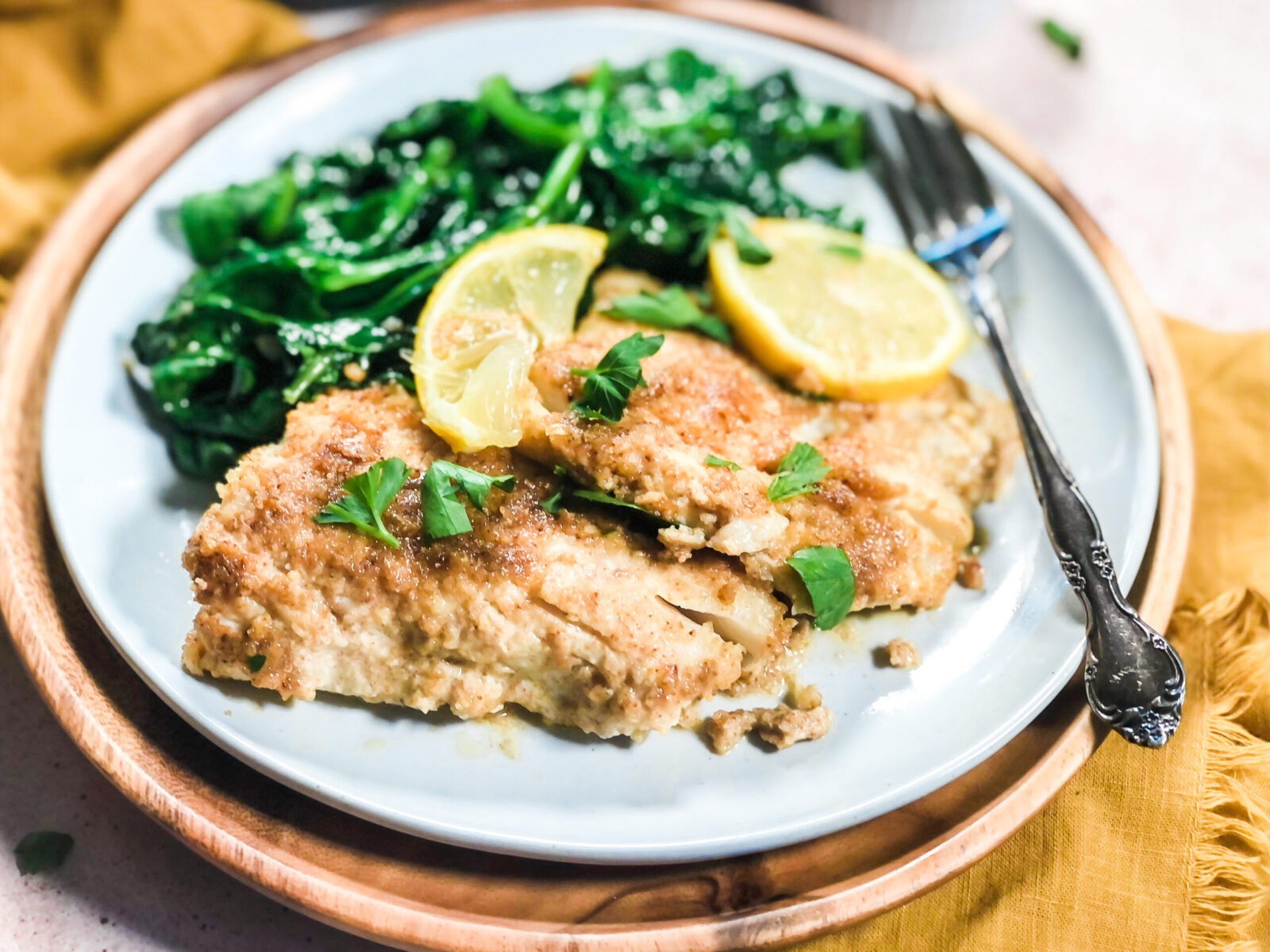 A plate of baked cod garnished with chopped fresh parsley and two lemon slices, with sautéed spinach and a silver fork, placed on a wooden charger plate.