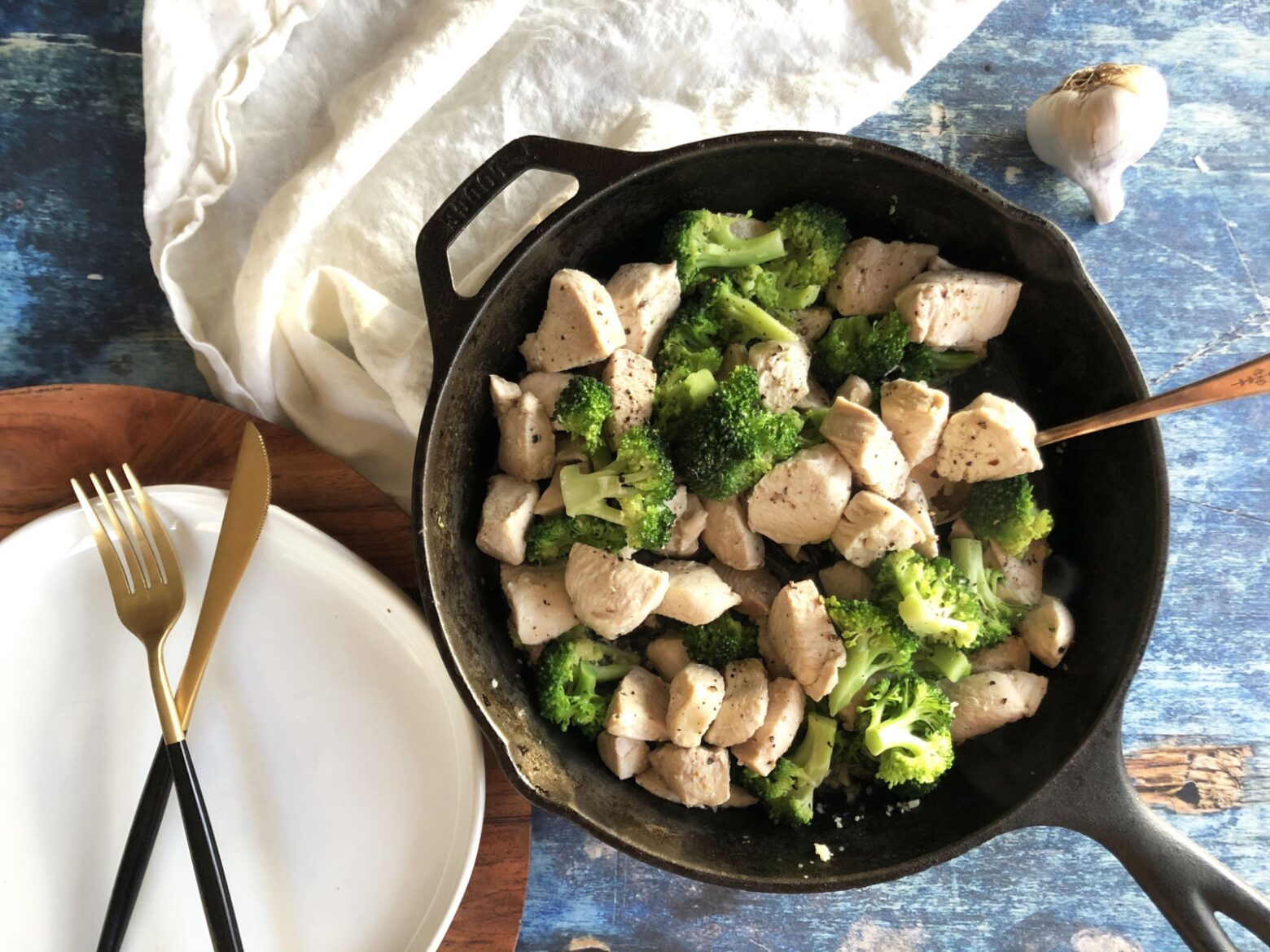 An iron skillet containing stir-fried chicken and broccoli next to an empty white plate with a knife and fork and a bulb of garlic.