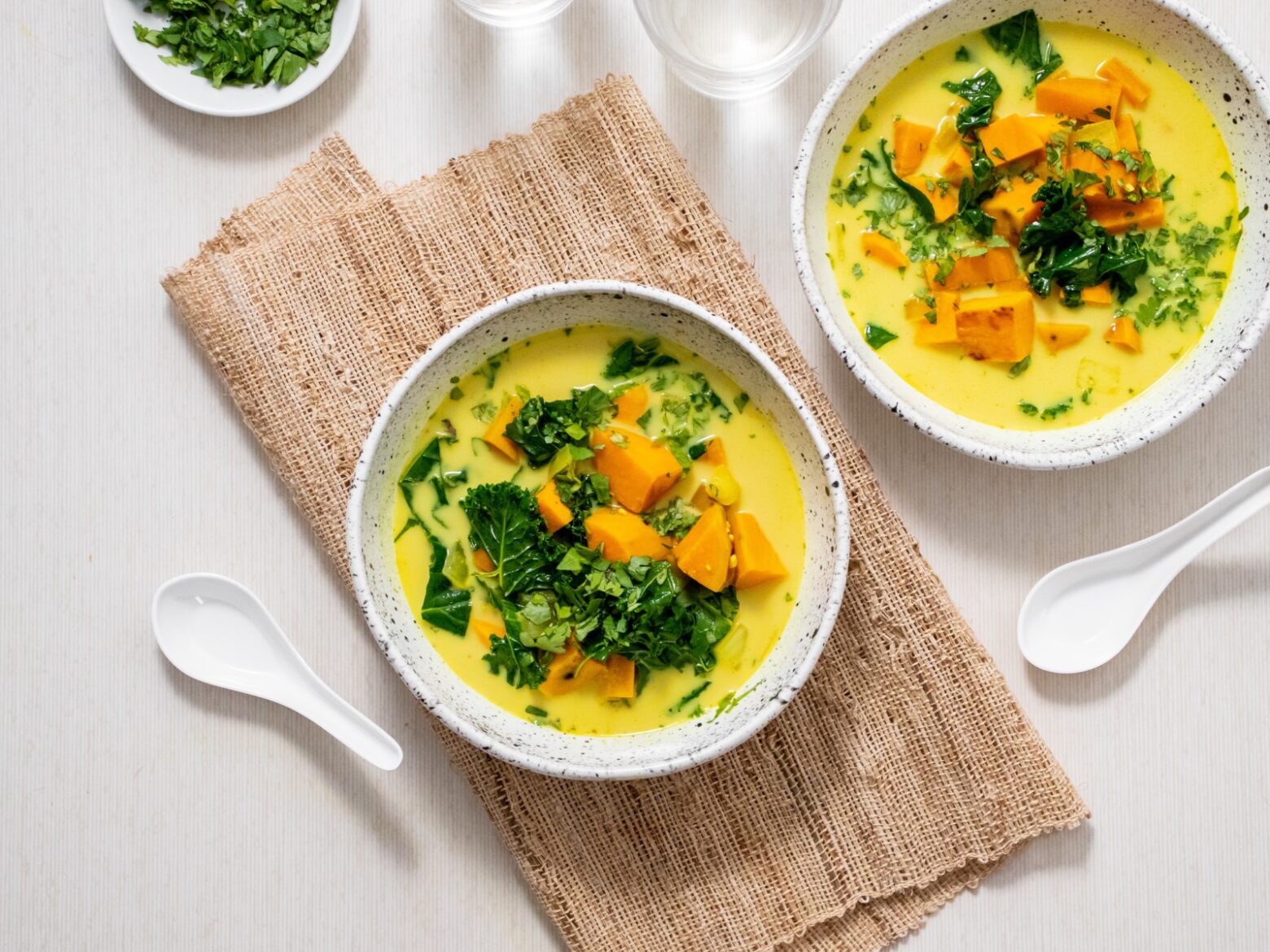 From above, two bowls of sweet potato turmeric soup, one on a folded burlap table cloth, surrounded by a small bowl of chopped cilantro and two white soup spoons.