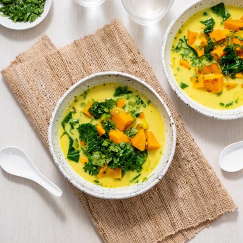 From above, two bowls of sweet potato turmeric soup, one on a folded burlap table cloth, surrounded by a small bowl of chopped cilantro and two white soup spoons.