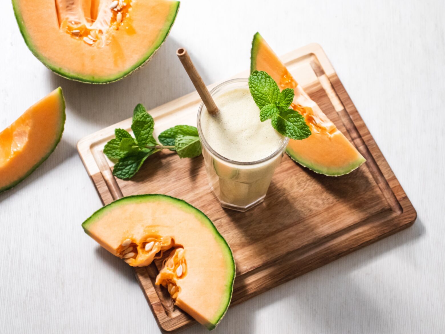 From above, a glass of cantaloupe ginger smoothie garnished with mint leaves on a wooden chopping board, surrounded by mint leaves and slices of cantaloupe.