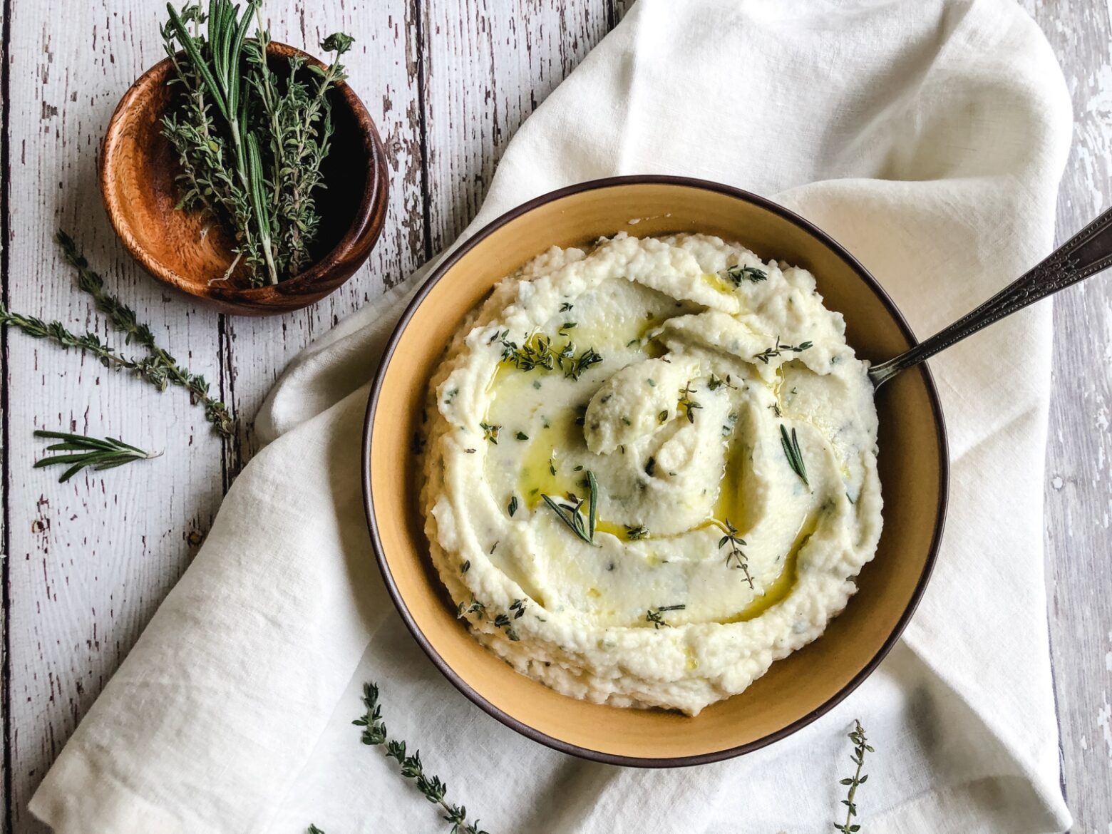 From above, a bowl of garlic mashed cauliflower garnished with chopped herbs next to a small wooden bowl of thyme, rosemary, and sage.