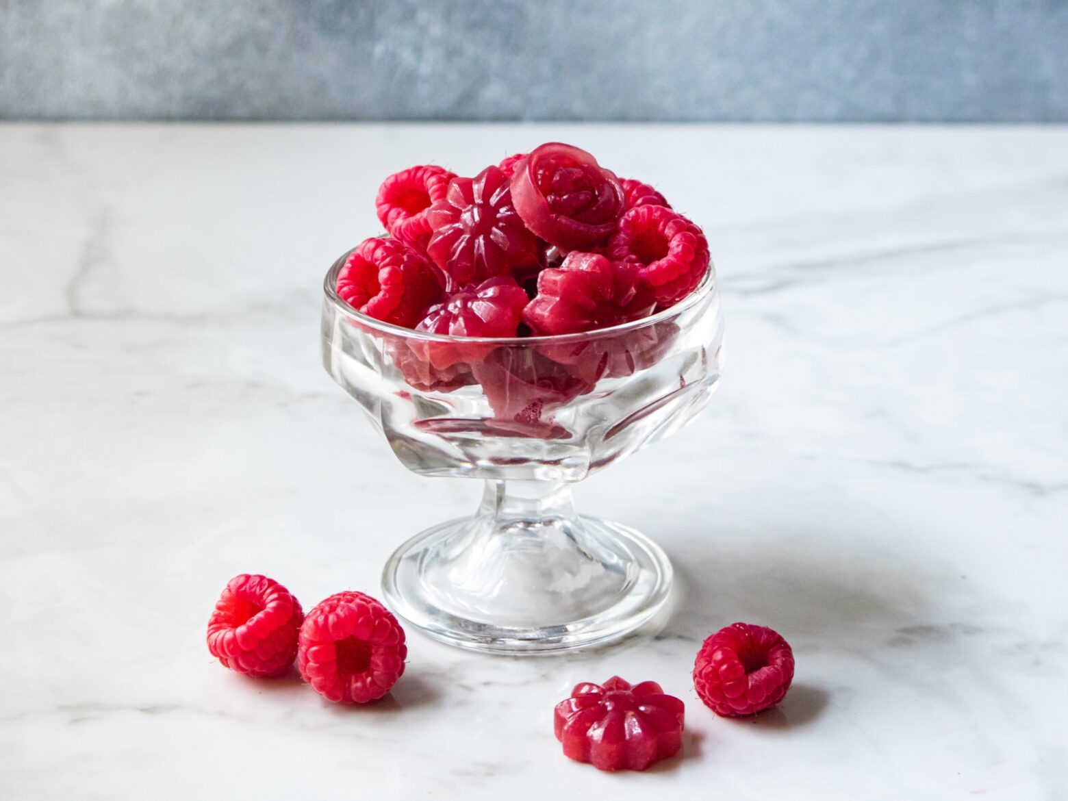 A small glass desert bowl filled with raspberries and pomegranate raspberry gummies, with three raspberries and one gummy on the white marble table next to the glass.