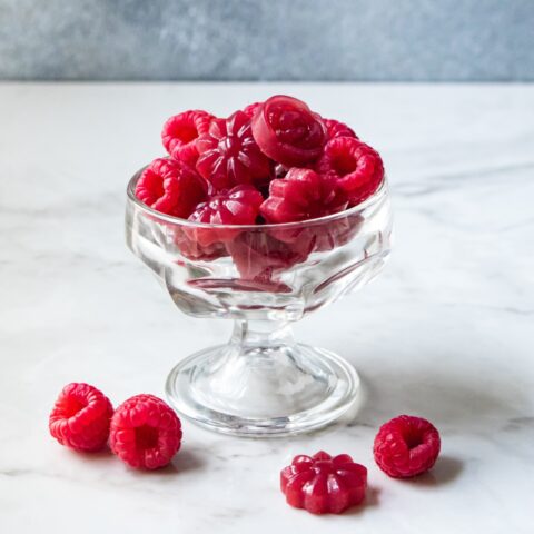 A small glass desert bowl filled with raspberries and pomegranate raspberry gummies, with three raspberries and one gummy on the white marble table next to the glass.