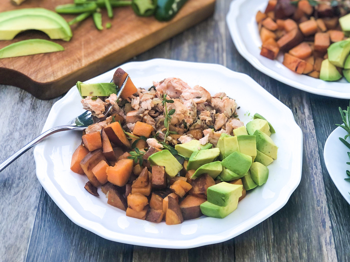Two bowls of spicy salmon with avocado and sweet potatoes, next to a wooden chopping board with sliced avocado and jalapeño.