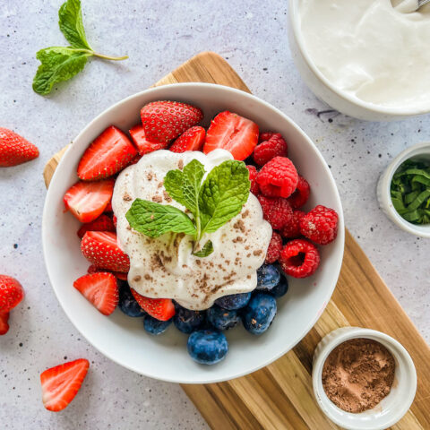 A bowl of raspberries, blueberries, and sliced strawberries, topped with coconut cream sprinkled with cacao powder, and fresh mint leaves.