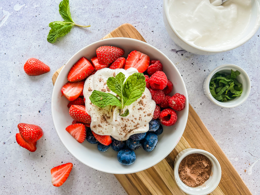 A bowl of raspberries, blueberries, and sliced strawberries, topped with coconut cream sprinkled with cacao powder, and fresh mint leaves.