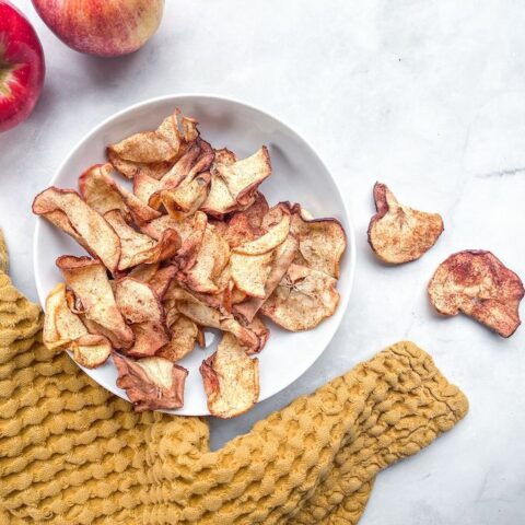 From above, a white bowl of spiced apple chips next to a tea towel, two apples and three apple chips.
