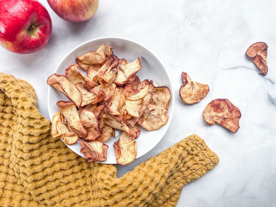 From above, a white bowl of spiced apple chips next to a tea towel, two apples and three apple chips.
