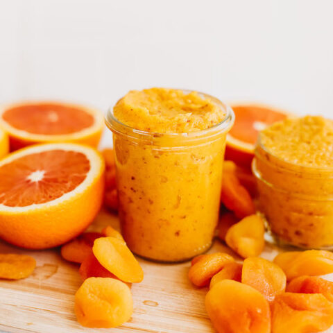 Two different sized glass jars of apricot jam on a wooden cutting board, surrounded by dried apricots and orange halves.