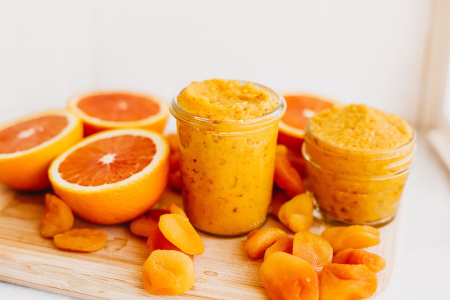Two different sized glass jars of apricot jam on a wooden cutting board, surrounded by dried apricots and orange halves.