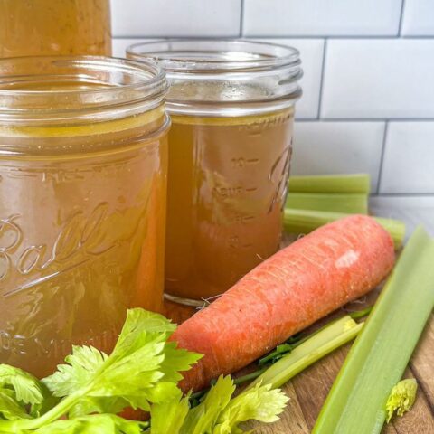 Three mason jars filled with chicken bone broth on a wooden tray surrounded by celery sticks and a carrot.