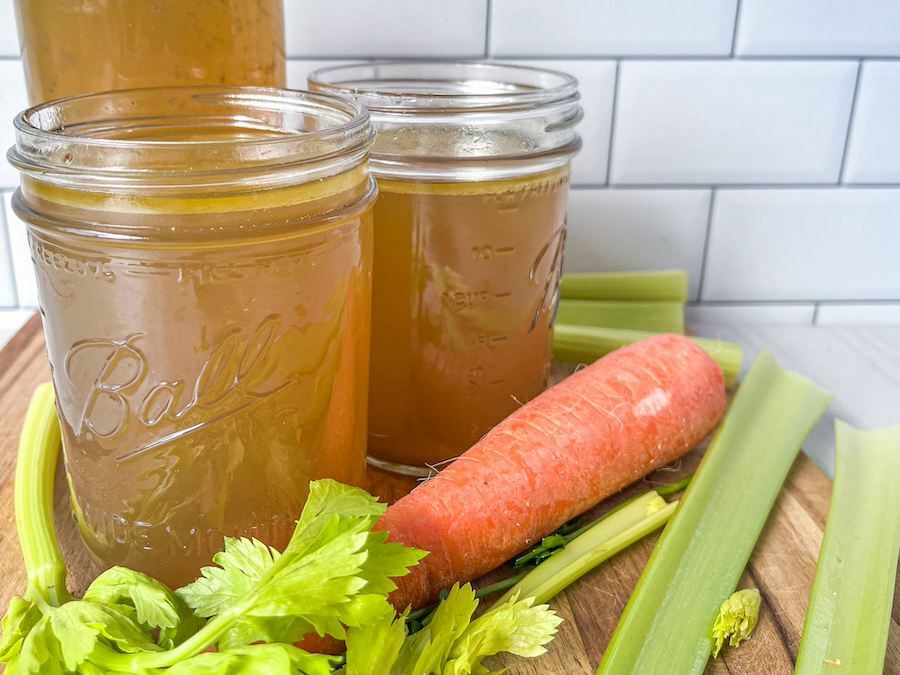 Three mason jars filled with chicken bone broth on a wooden tray surrounded by celery sticks and a carrot.