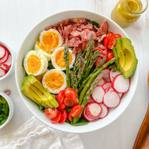 From above, a white bowl full of a Paleo compliant Cobb salad, surrounded by small bowls of sliced radishes, chopped green onions, a jar of mustard and two wooden spoons.