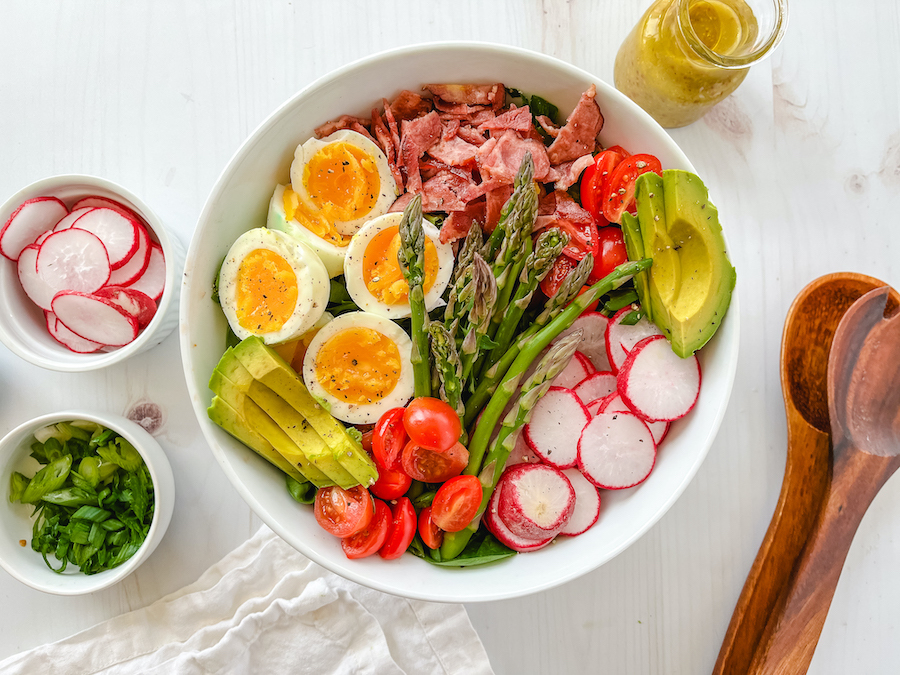From above, a white bowl full of a Paleo compliant Cobb salad, surrounded by small bowls of sliced radishes, chopped green onions, a jar of mustard and two wooden spoons.