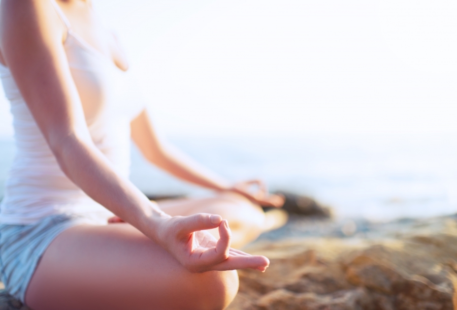 A woman in a white tank top shirt sits on a beach in a tradition post of meditation.