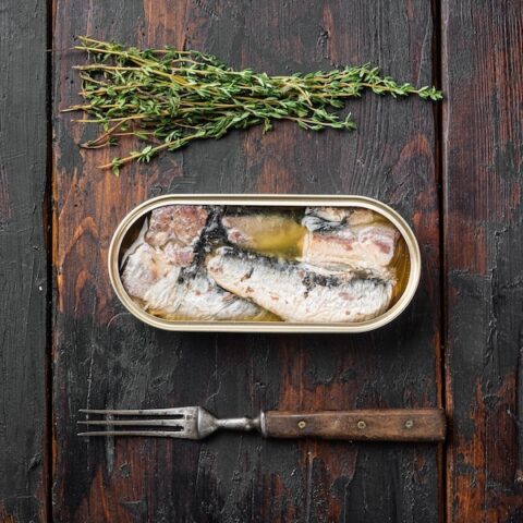 Tin of canned sardines with a fork and some herbs on a table.