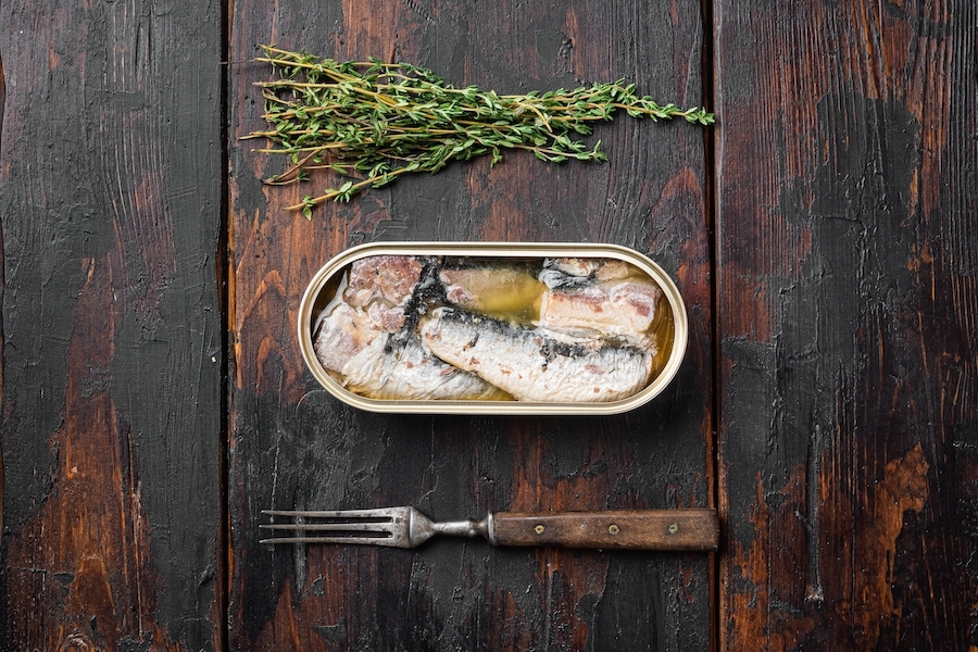 Tin of canned sardines with a fork and some herbs on a table.