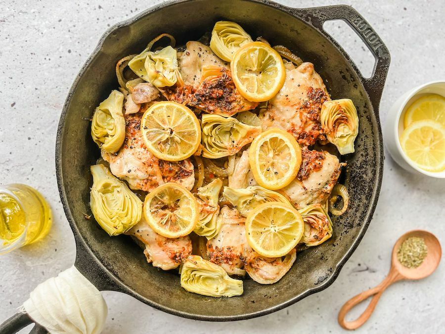 A cooked dish of chicken, artichoke hearts, and lemon slices in an iron skillet.
