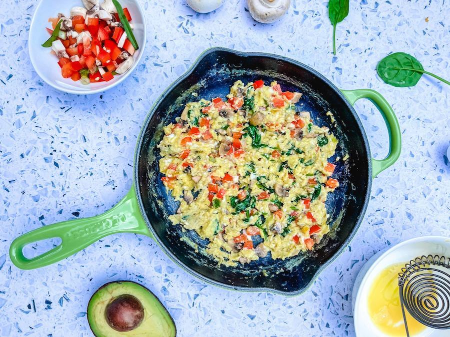 From above, an iron skillet with scrambled eggs and vegetables, surrounded by a bowl of chopped uncooked vegetables, two raw mushrooms, spinach leaves, half an avocado and a cracked raw egg in a bowl.
