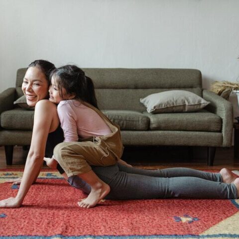 A woman smiles while doing a yoga pose with her young child on her back.