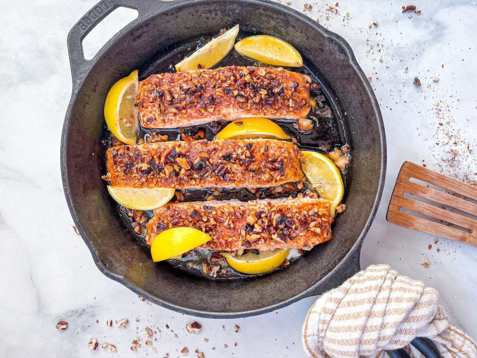 From above, an iron skillet with three pecan crusted salmon fillet slices surrounded by lemon wedges.