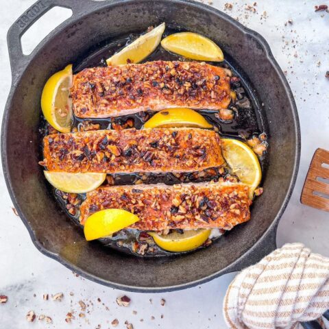From above, an iron skillet with three pecan crusted salmon fillet slices surrounded by lemon wedges.
