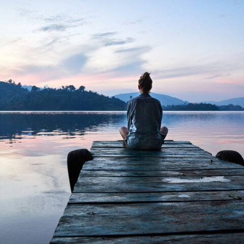 A woman sits on the end a pier in front of a calm lake with mountains behind at sunrise.