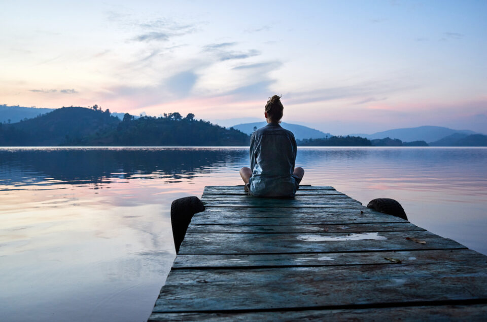 A woman sits on the end a pier in front of a calm lake with mountains behind at sunrise.