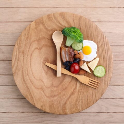 A wooden plate is set with utensils in the shape of a clock with food inside the utensils to indicate an intermittent fasting schedule.