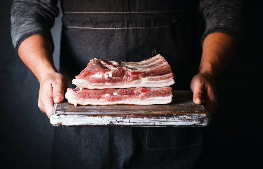 Man in an apron holding a cutting board with two slabs of pork ribs on it.