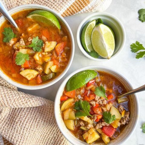 From above, two bowls of taco soup both with silver spoons and a lime wedge and garnished with cilantro, next to a small bowl of lime wedges and cilantro leaves.