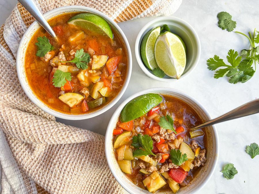 From above, two bowls of taco soup both with silver spoons and a lime wedge and garnished with cilantro, next to a small bowl of lime wedges and cilantro leaves.