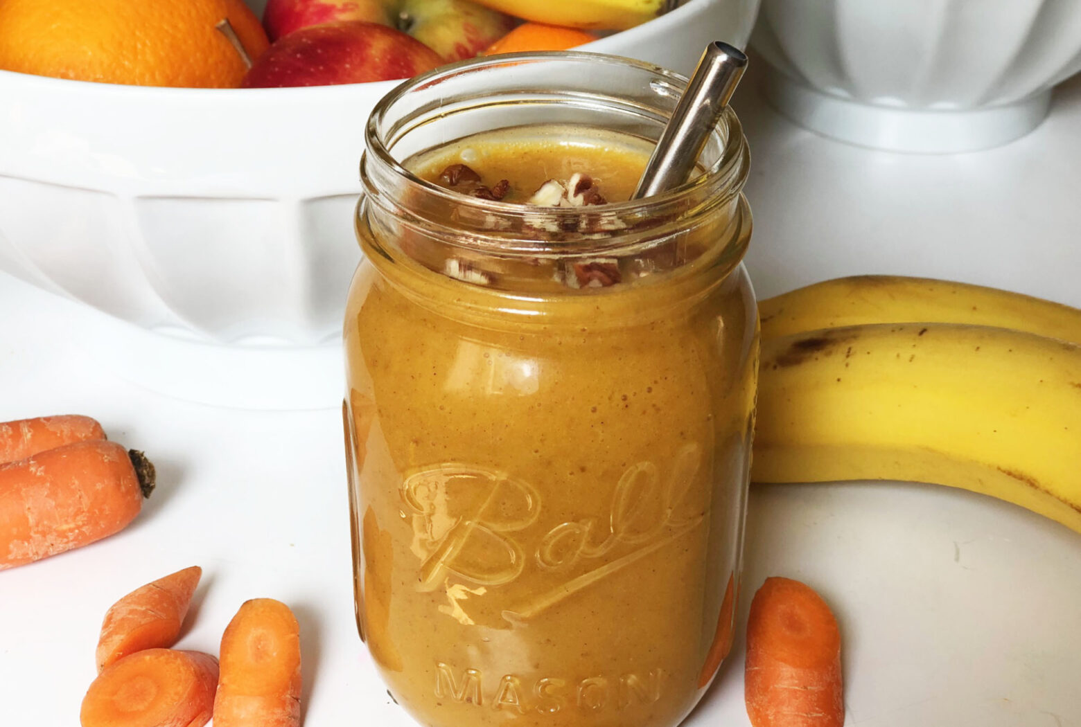A mason jar full of pumpkin pie smoothie with a silver metal straw, garnished with chopped pecans and surrounded by a large white bowl of fruit, two bananas and some carrots.