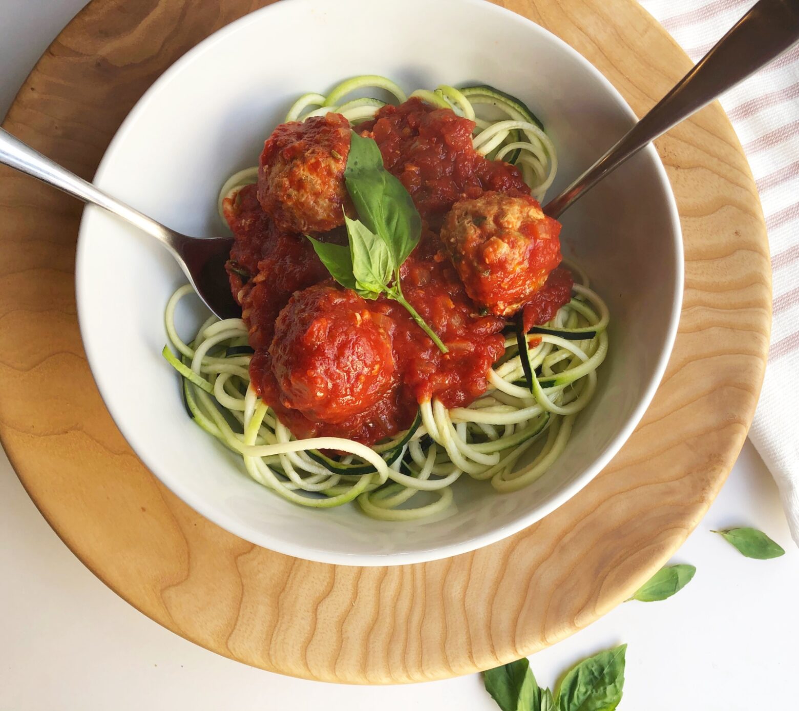 From above, on a wooden charger, a white bowl of zoodles and turkey meatballs with a silver fork and spoon, garnished with fresh basil.